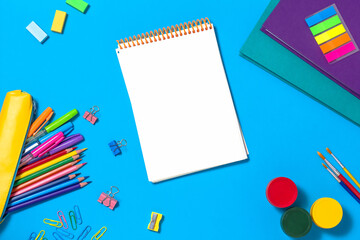 The concept of going back to school. Colorful school supplies, notebooks, pencils, pens, paper clips, paint on a blue background. Space for text. The view from the top