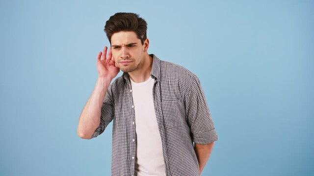 Young bunet male in casual clothes is trying to hear something easily, but he doesnt manage. Posing against blue studio background