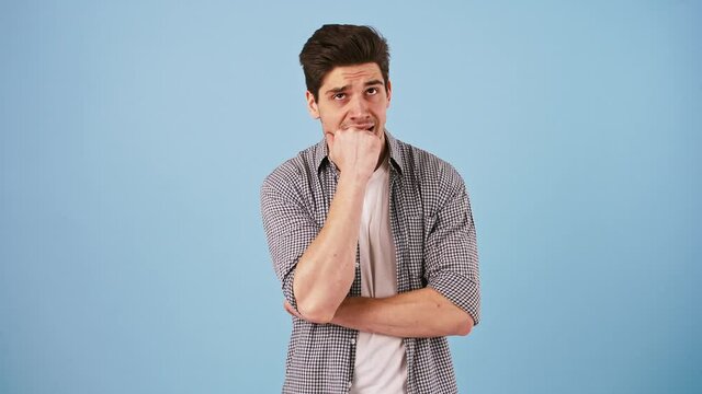 Young bunet guy in casual outfit is biting his nails and fist, looking worried, nervous or scared. Posing against blue studio background
