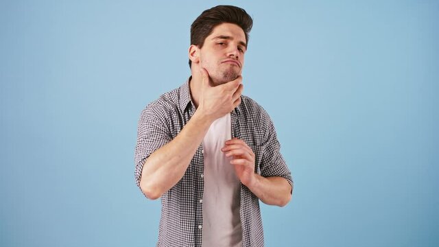 Young man acting like looking into mirror. Straightening hair and eyebrows, looking at face, smiling, pointing at himself. Posing on blue background