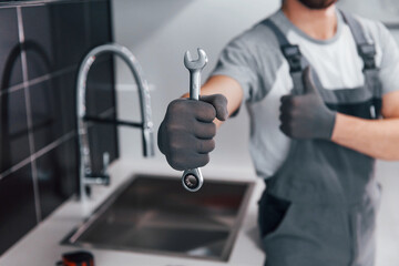 Close up view of young professional plumber in grey uniform holding wrench in hand on the kitchen