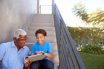 Grandfather With Grandson Sitting On Steps Outdoors At Home Using Digital Tablet