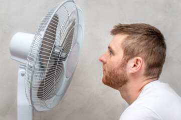 young man stands by the fan. Close-up portrait. The man is hot in the apartment. Hot weather concept