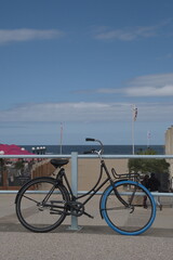 Fototapeta na wymiar Ein schwarzes Fahrrad mit einem blauen Rad vorne und Edelstahl Griffe steht geparkt am Ufer des Meeres. Der Himmel ist hell blau ,das Meer dunkel, der Sand und der Boden des Ufers sind hell.