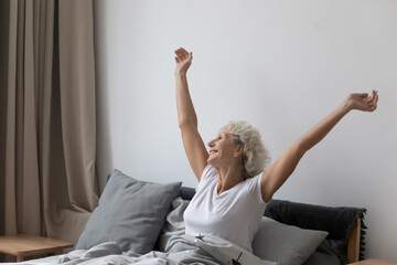 Optimistic refreshed middle aged senior woman sitting in bed wake up feel rested after enough healthy sleep in comfortable bed, stretching arms smiling looking out the window enjoy and welcome new day