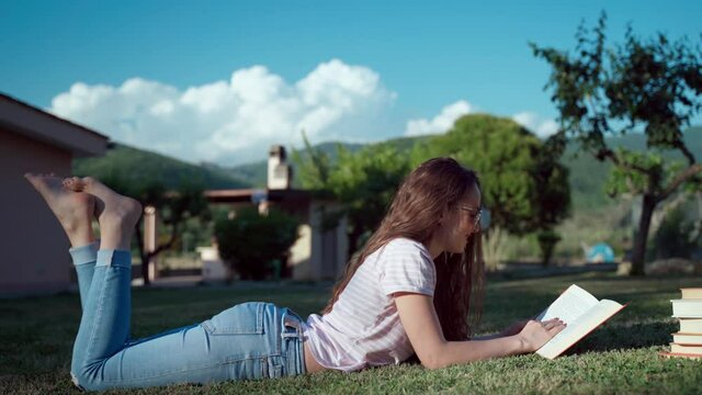 Pretty girl in sunglasses lying on the grass near the house on sunset background and reading interesting book, long brown curly hair girl relaxing outdoors among the piles of books, knowledge and