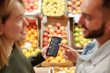 Positive young couple standing at food counter and using smartphone app while discussing product list at organic market