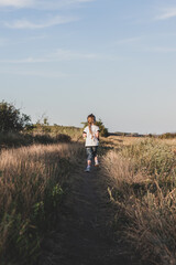 Naklejka na ściany i meble Adorable little girl with long braid running down the hill back view. Happy child running in wild grass countryside landscape. Family walking on fresh air. Local hiking travel concept.