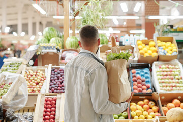 Rear view of young man in light denim jacket holding paper bag and choosing juicy fruits at farmers market