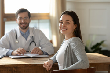 Portrait of young beautiful smiling female client patient with blurred male general practitioner surgeon on background. Happy woman visiting family doctor, enjoying checkup meeting in hospital.