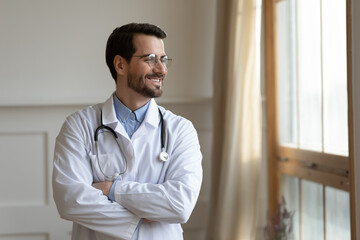 Smiling young doctor in white medical uniform standing with folded hands near window, lost in thoughts. Positive handsome male head of department thinking of challenges, planning future career.