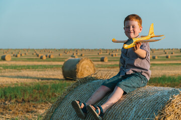 Naklejka na ściany i meble The red-haired boy plays with a toy airplane on a wheat field with bales