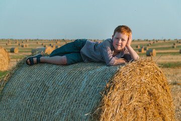 Naklejka na ściany i meble The red-haired boy lies on top of a straw bale on a wheat field