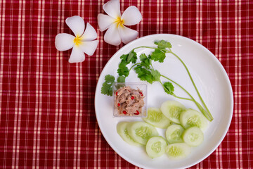 Spicy tuna salad in a small plastic cup placed on white plate, sliced ​​cucumber and coriander. Food is laid out on red background with scotch pattern, side of plate has plumeria flowers.