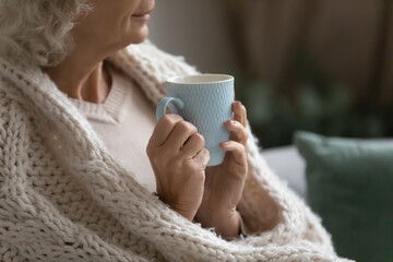 Aged calm woman resting wrapped in knitted plaid drink evening tea look into distance, close up....