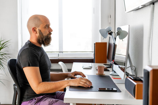 Hombre calvo con camiseta gris oscura, pantalón corto púrpura, y barba, trabajando en su ordenador con pantalla en blanco, en su oficina, iluminado por luz natural