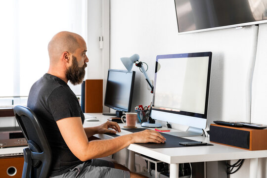 Hombre calvo con camiseta gris oscura, pantalón corto púrpura, y barba, trabajando en su ordenador con pantalla en blanco, en su oficina, iluminado por luz natural