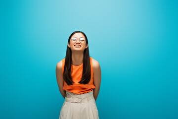 cheerful asian girl in glasses smiling and standing on blue