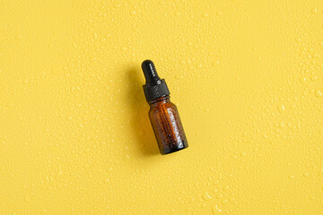 Brown amber glass cosmetic dropper bottle in droplets of water on yellow background.