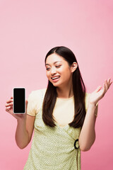excited asian woman looking at smartphone with blank screen isolated on pink