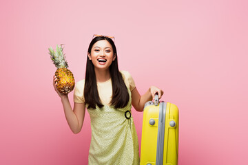 happy asian girl holding baggage and fresh pineapple isolated on pink