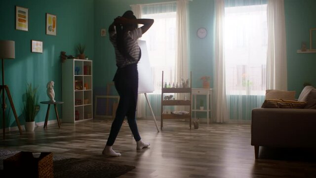 Pretty black woman enters a room, takes a brush, is going to paint on big white canvas, being inspired to create in her cozy apartment, creative atmosphere and mood, Slow motion.