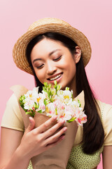 young asian woman in straw hat smiling and holding flowers isolated on pink