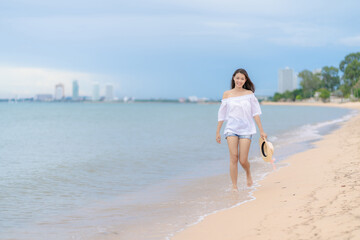 Fototapeta na wymiar Happy traveler asian woman in white casual wear holding a hat while enjoying a walk on the sea beach at Pattaya, Thailand. Summer travel and vacation concept,