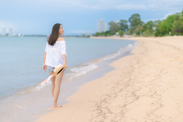 Happy traveler asian woman in white casual wear holding a hat while enjoying a walk on the sea beach at Pattaya, Thailand. Summer travel and vacation concept,