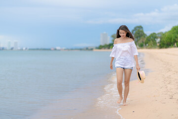Fototapeta na wymiar Happy traveler asian woman in white casual wear holding a hat while enjoying a walk on the sea beach at Pattaya, Thailand. Summer travel and vacation concept,