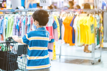 Back view of boy wearing face mask to prevent the spread of the Corona Virus (Covid-19) shopping in a supermarket. Boy with shopping cart during virus outbreak in grocery store. New normal lifestyle.