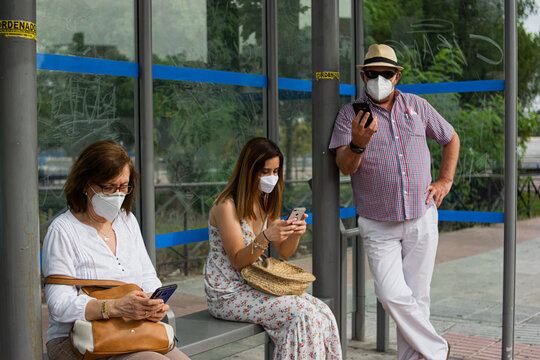 Photo of a three people waiting for the bus checking her phone in a bus stop wearing a white face mask and keeping social distance