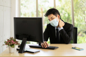 A young Asian businessman is wearing a suit wearing a face mask looking exhausted while sitting at his working place sitting at an office desk in the office.Feeling exhausted.