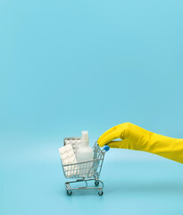 Fototapeta premium Female hand in a yellow medical glove holds a toy shopping cart with medicines on a blue background. Hygiene in the store. Coronavirus pandemic, covid-19.