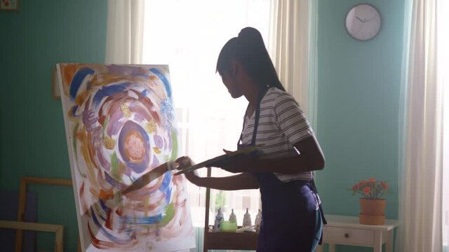 Young black woman student entertains at home dancing and painting on big canvas, choosing bright saturated colors, transmits inner energy into new project, feeling free and happy, Slow motion.
