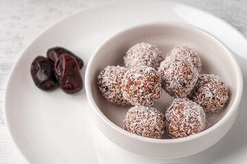 Candy balls made of dates and coconut shavings in a white dish on a light background near the dates