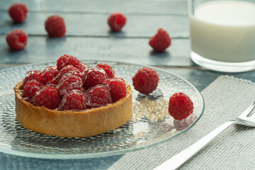 Raspberry tartlets served on a transparent plate with brown sugar. Glass of milk on a light blue wooden background.