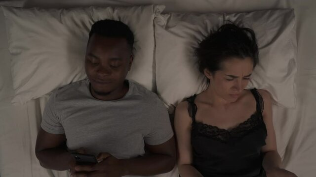 Mixed ethnicity sleepless couple lying in bed at night. White woman pinching nose, doing bad smell gesture. Black man holding phone in hands and smiling