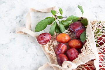 A bunch of ripe plums and a twig with leaves lie in a reusable mesh bag on a light concrete background. Fruit still life with copy space for text. Top view.