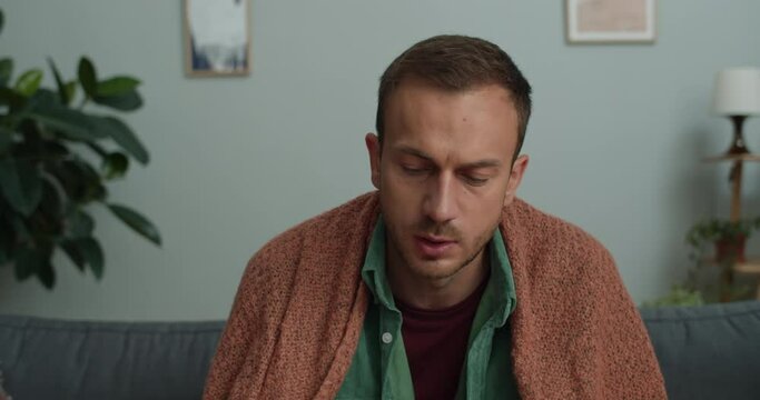 Bearded young man talking and showing medicine while looking to camera. Ill guy in 40s caughing while having online conversation with doctor and sitting on sofa at home