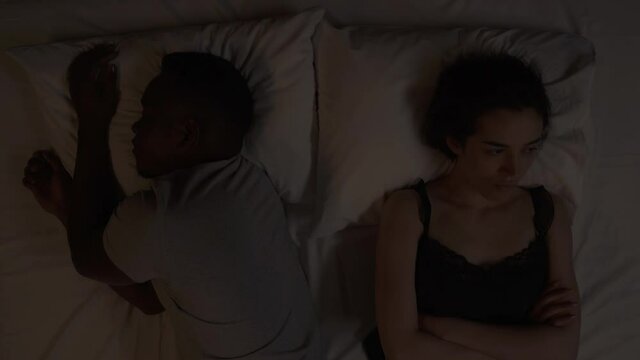 Close-up of multiracial couple lying in bed at night. Caucasian girl sights and shaking head in irritation because her African boyfriend is sleeping