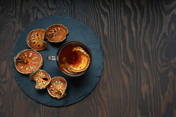 Top view of thai healing matoom tea made of dried bael fruit slices, also known as wood apple, in glass cup on round stony tray on dark wooden background. Image with copy space