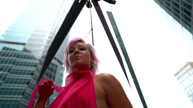 A close-up portrait of an informal woman with light pink hair is on the street during the day against the background of glass high-rises and business centers. She's wearing a bright pink dress