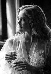 Black and white portrait of elegant bride sitting in front of window holding glass of champagne.
