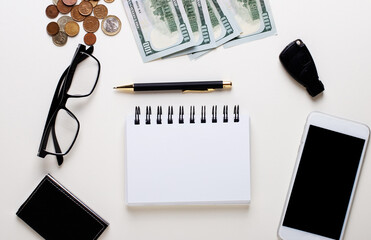 On a light steel sheet lie money, a car key, a smartphone, a business card holder, black-framed glasses and a blank white notebook. Copy space. Business concept