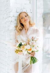 Fototapeta premium The blonde in a gentle peignoir stands by the window with a bouquet in her hands.