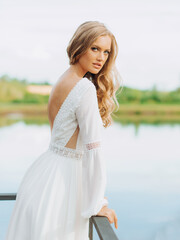 Fototapeta na wymiar Blonde hair bride in white silk long lace dress standing on the terrace with lake view