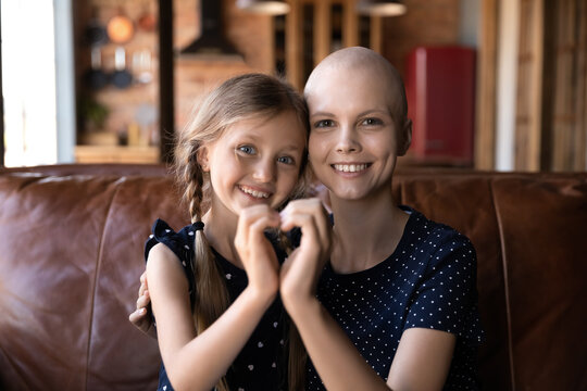 Portrait of smiling young sick of cancer bald mom and little daughter hug show heart love sign gesture with hands, happy hopeful ill hairless mother patient with small girl child, healthcare concept