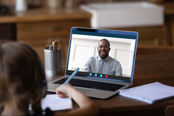 Back view of little schoolgirl sit at table look at laptop screen have online video call lesson with biracial male teacher or tutor, small girl child study on webcam conference with coach on computer