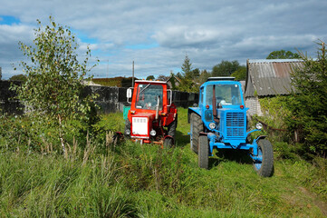 Fototapeta premium Two tractors in a village near the wood house. Red and blue tractor are in the yard.